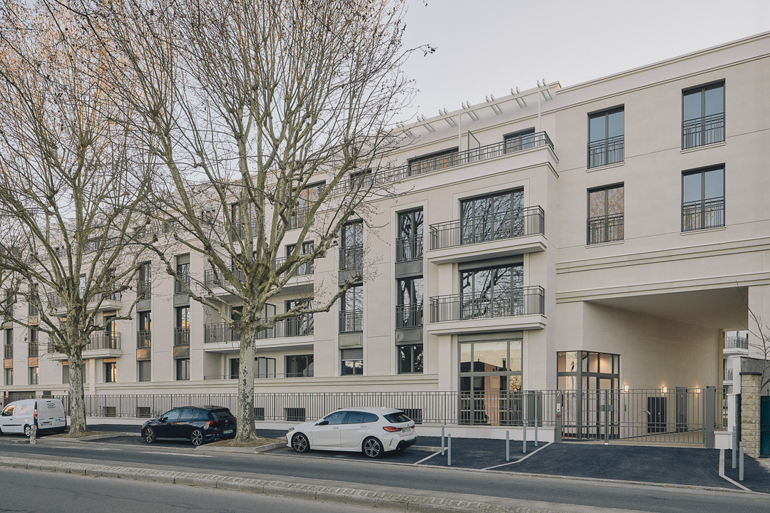 Résidence contemporaine de logements collectifs, composée de volumes blancs aux lignes sobres, le long d'un boulevard, en lumière de fin de journée.
